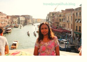 A travel blogger in a floral pink dress standing on the Rialto Bridge, with a scenic view of the Grand Canal, gondolas, and historic Venetian architecture in the background. Featured on traveljournalhub.blog